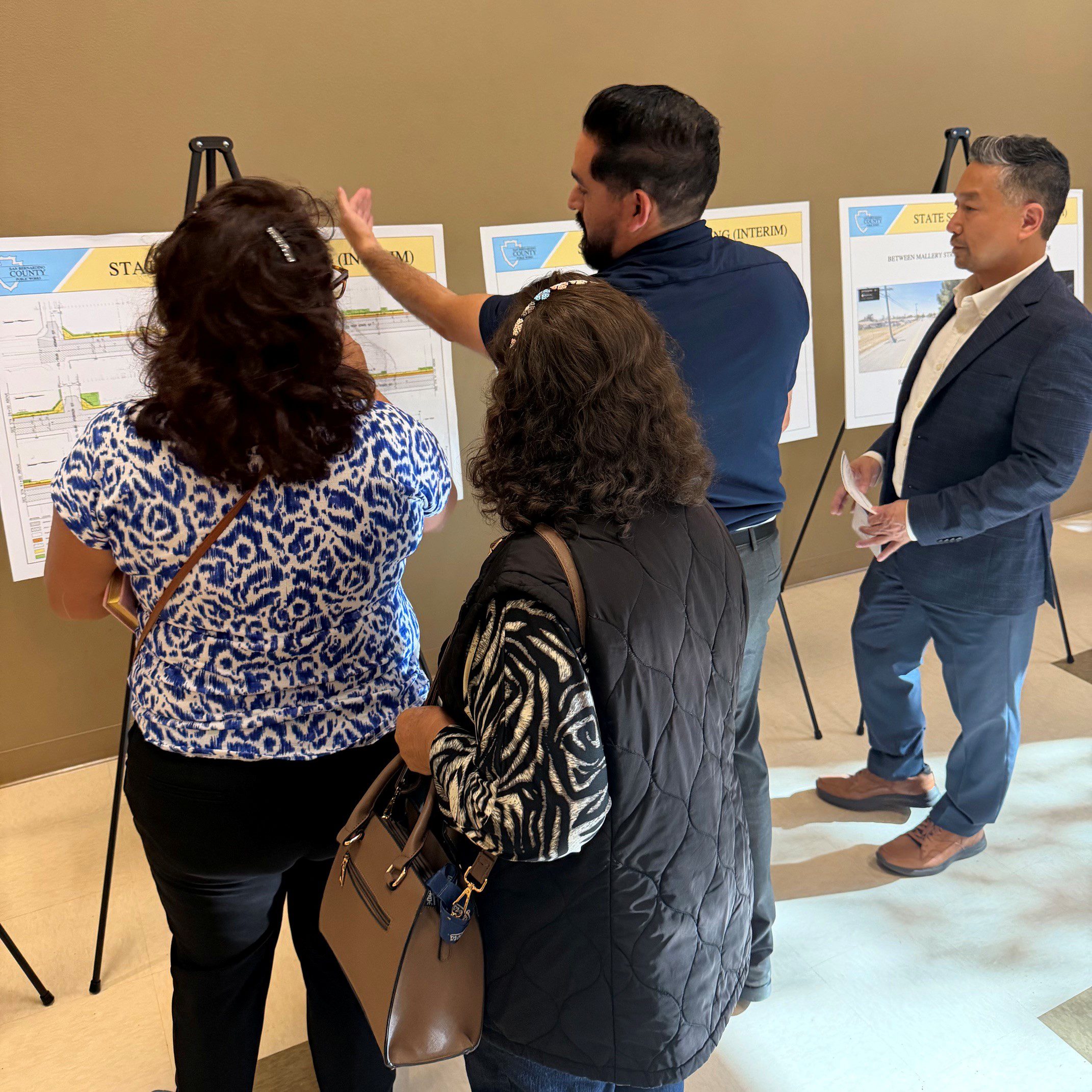 Group reviews roadway design posters on easels while a presenter gestures during a San Bernardino County Public Works workshop.