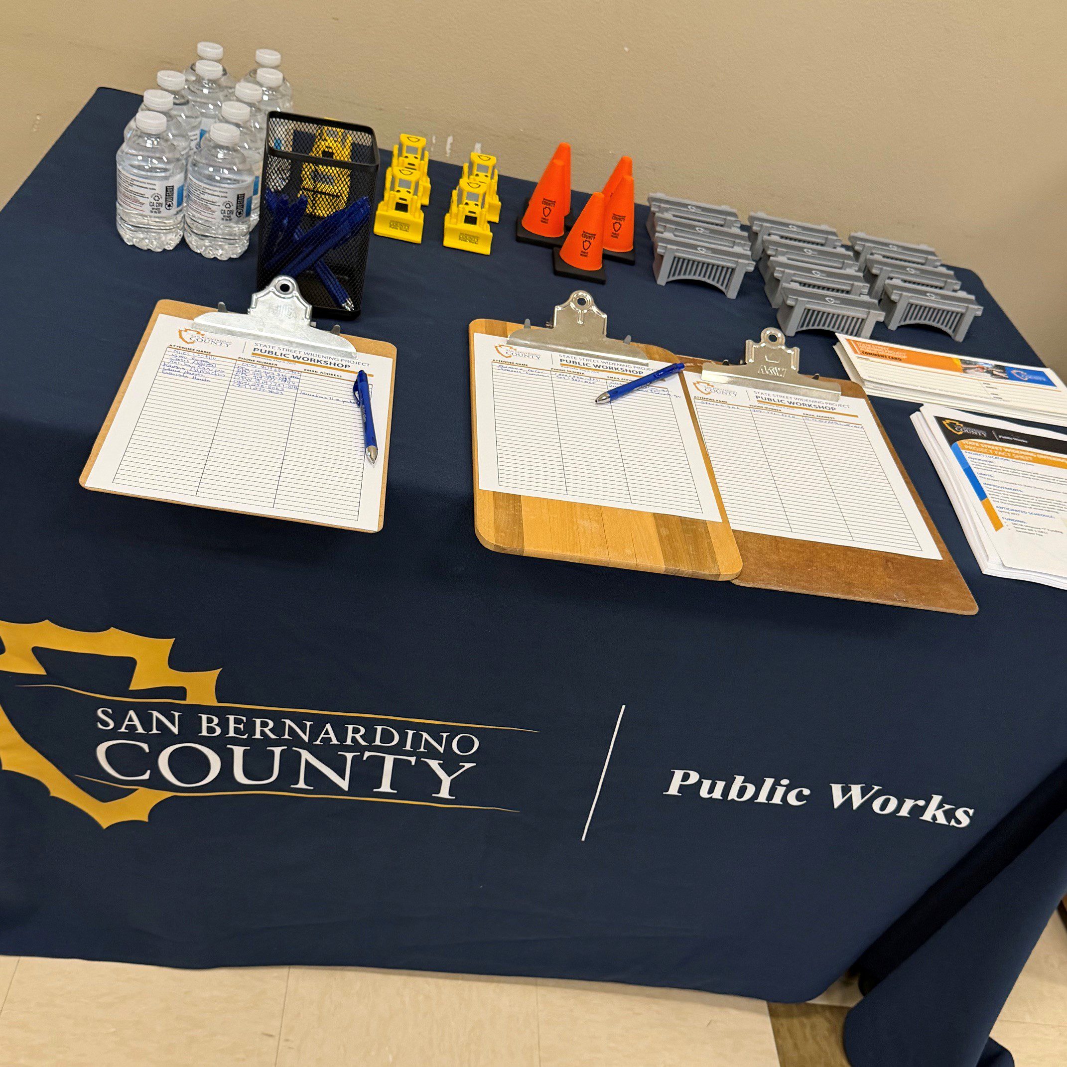 Table with San Bernardino County Public Works sign-in sheets, pens, water bottles, brochures, and small traffic demo props.
