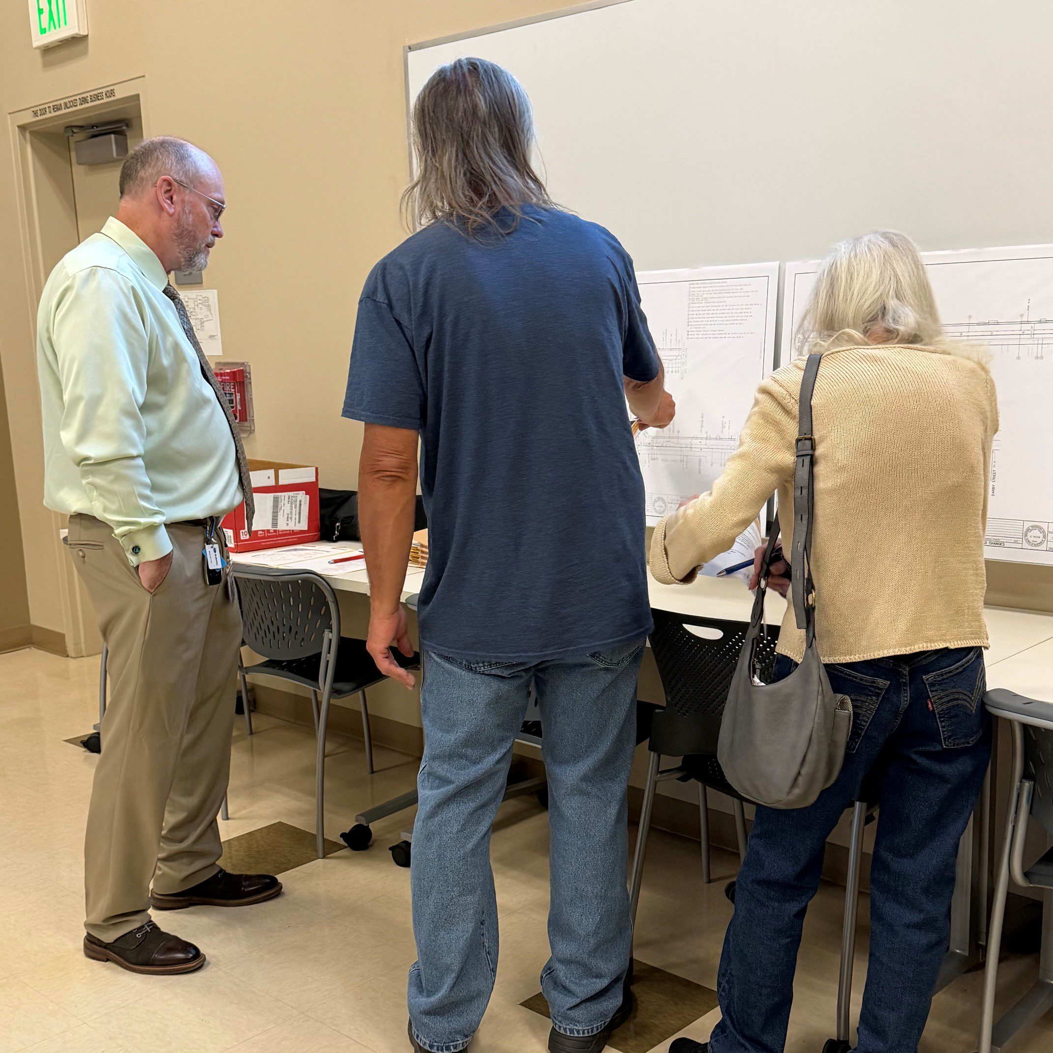 Three people examine engineering plans on a table, discussing details during a public works project review.